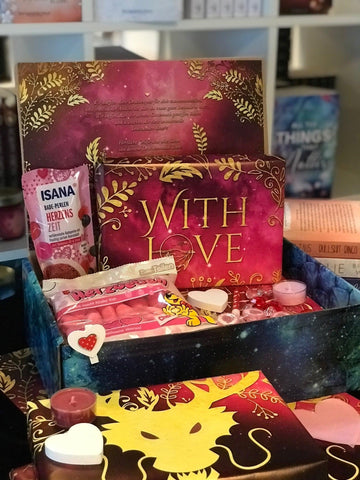 Das Produktbild zeigt vorn eine geöffnete Drachenpost in blauem Schuppen-Design. Man sieht den in den Boxdeckel geklebten Brief zum Thema #withlove und in der Box ein in ein #withlove-Poster verpacktes Buch und zum Thema passende Goodies - Süßigkeiten und andere Kleinigkeiten. Im Hintergrund sieht man die Bücherregale des Verlagsladens.